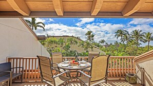 A covered outdoor dining area with a table set for four, surrounded by wicker chairs, overlooking a tropical landscape with palm trees and a clear sky.