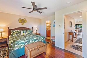 A bedroom with a bed, a ceiling fan, a nightstand with a lamp, a wicker basket, and a view of the dining area through an open door. - Fully Renovated Kihei 2BR Condo New AC Tropical Grounds Waterfall Pool (Kihei)
