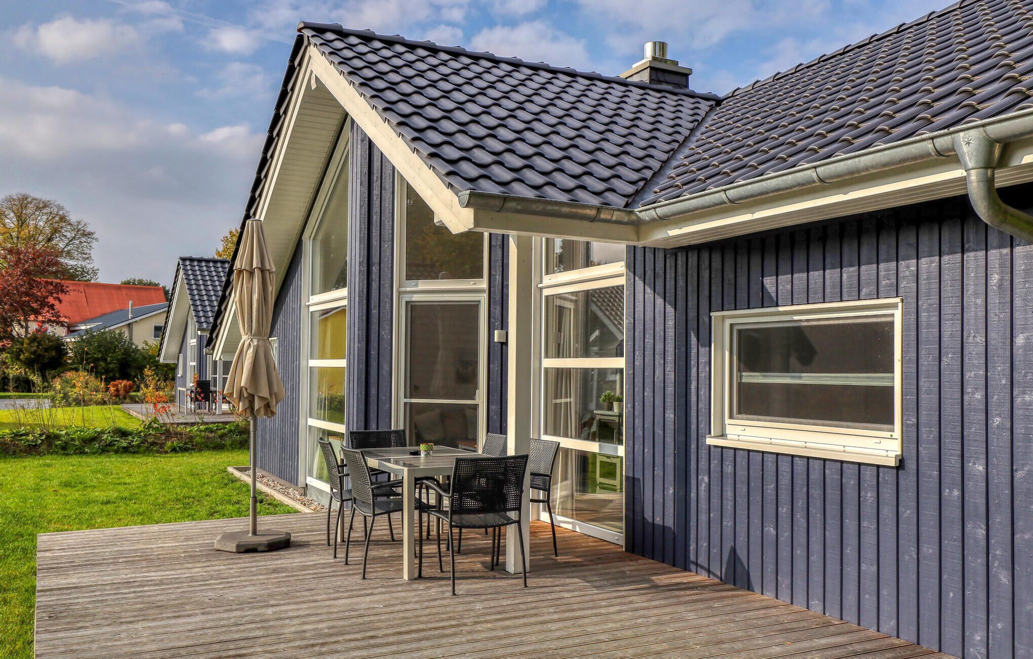 Una casa moderna con fachada azul oscuro, amplias ventanas y una terraza de madera con mobiliario exterior.