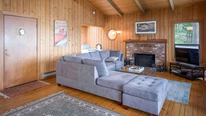 Un salon chaleureux avec un canapé d’angle gris, une cheminée et une grande fenêtre donnant sur l’extérieur.