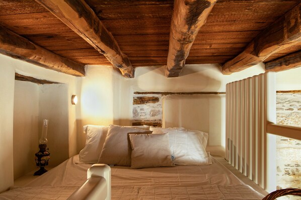 Une chambre avec un plafond en bois, un lit recouvert de draps blancs et une fenêtre équipée de stores.