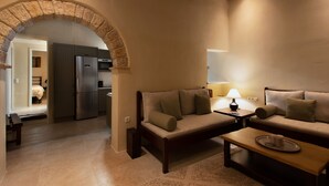 A cozy living area with a sofa, a coffee table, and a lamp. There is a kitchen visible in the background.