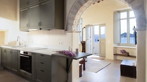 A kitchen with a large island, built-in oven, and grey cabinets. There is a stone archway leading to a balcony with a view.
