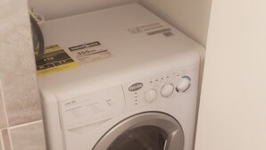 Een compacte, witte wasmachine met frontlader in een hoek van een kamer met betegelde vloer.
