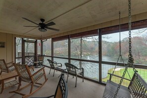 A covered outdoor area with a table, chairs, a rocking chair, a swing, and a view of a body of water and trees.