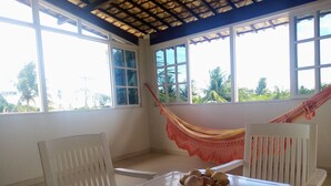 Ein Zimmer mit einer Hängematte, einem Tisch mit Obstschale und großen Fenstern mit Blick auf Palmen.