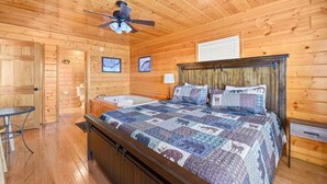 Un dormitorio tipo cabaña de madera con una cama grande, un ventilador de techo, una ventana y una mesita.