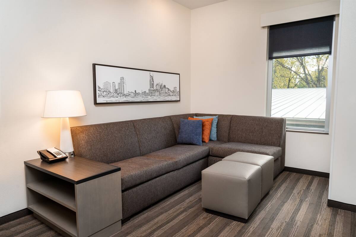 A modern hotel room with a grey sectional sofa, a small table with a lamp, a framed cityscape artwork, and a window with a view of trees.