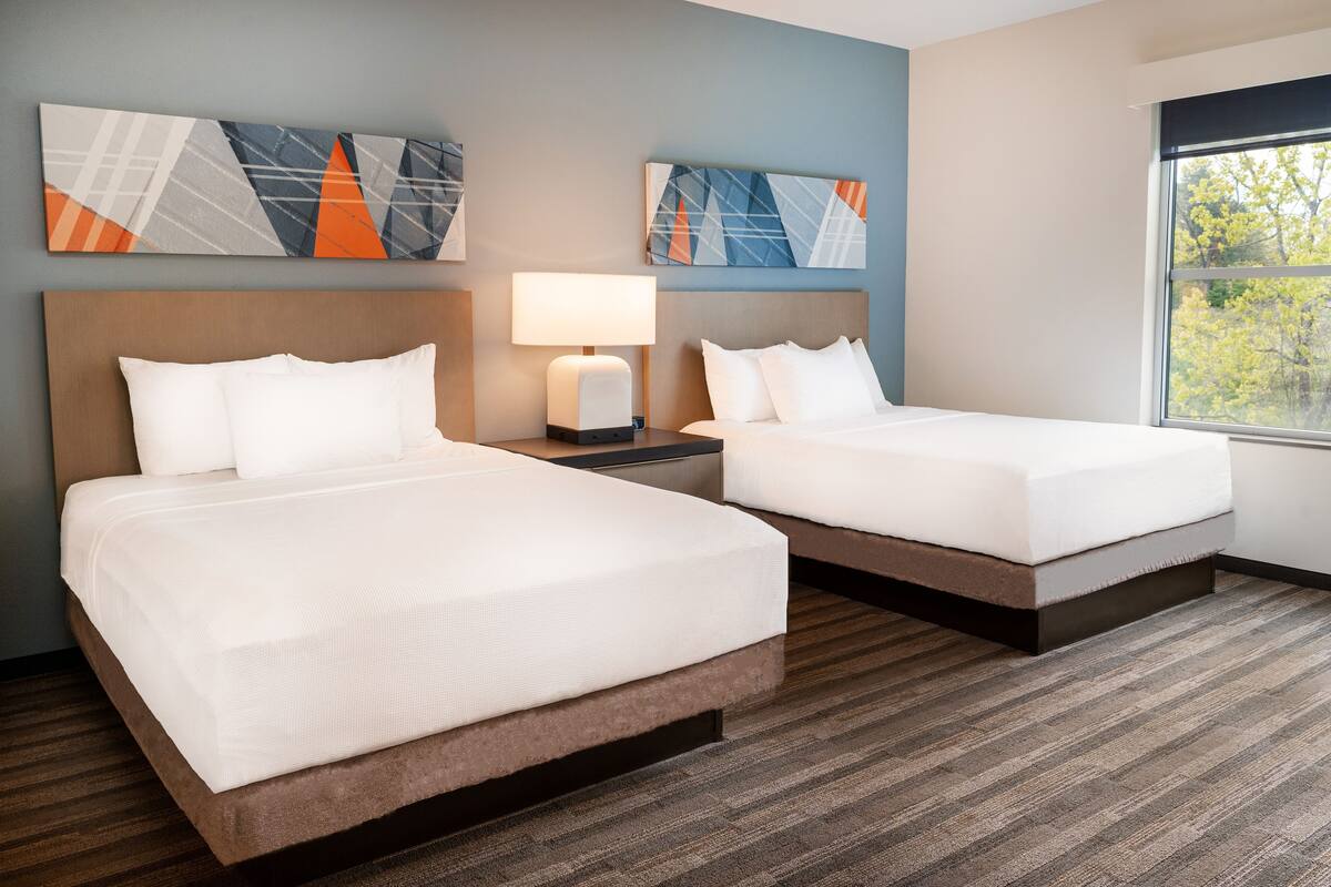 A hotel room with two beds, a nightstand with a lamp, abstract wall art, and a window with a view of trees.