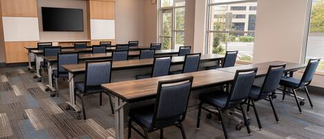 A conference room with a large table, chairs, a TV screen, and windows with a view of buildings and trees.