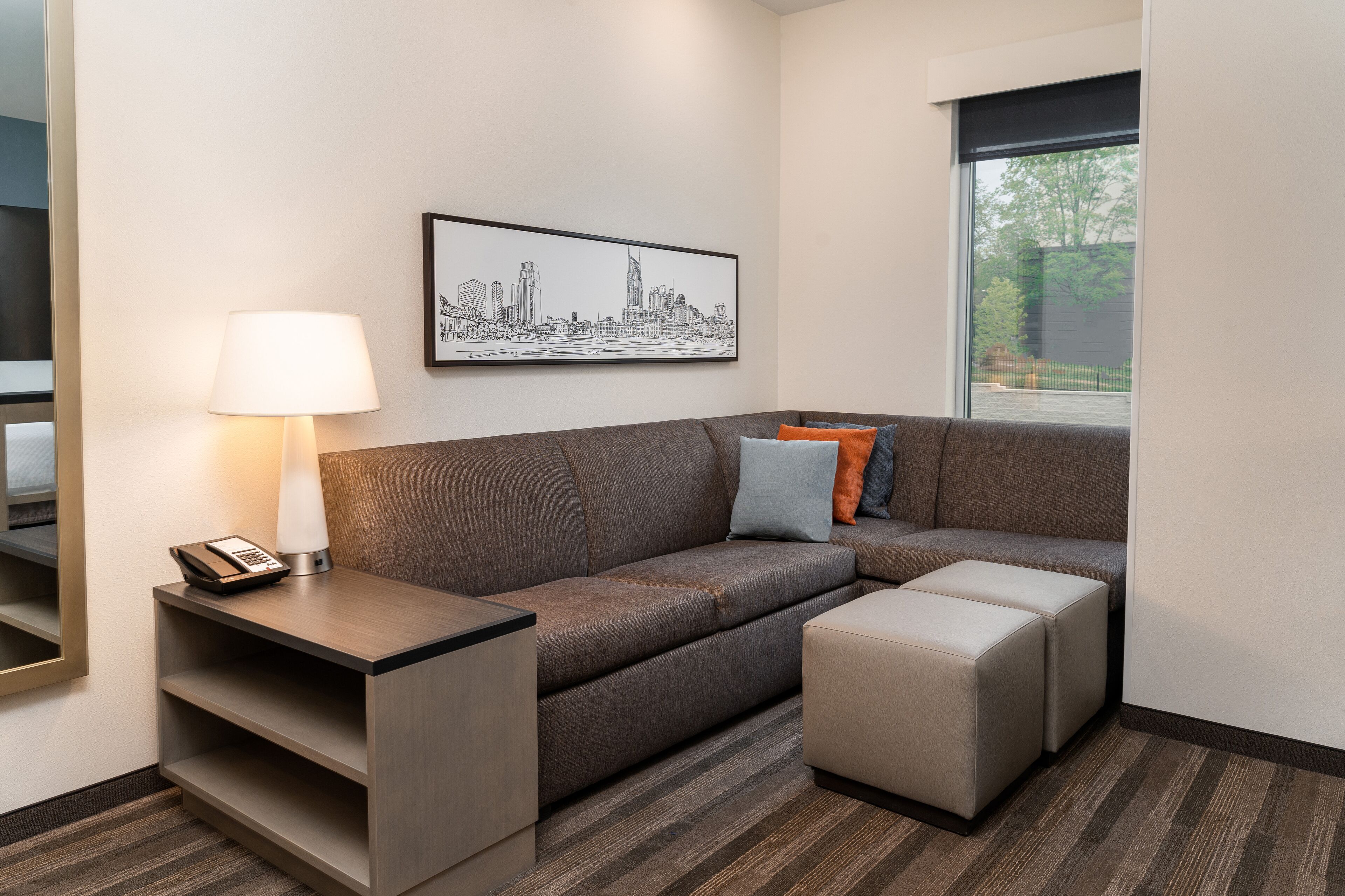 A modern hotel room with a gray sectional sofa, a small table with a lamp, and a framed cityscape artwork on the wall.