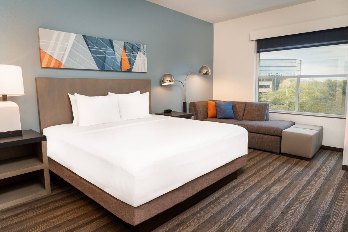 A hotel room with a large bed, a sofa, a desk, and a window with a city view.