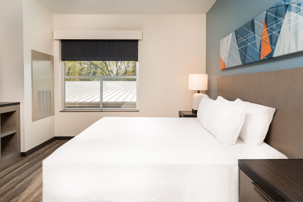 A hotel room with a large bed, a bedside table with a lamp, a window with a view of the outside, and a painting on the wall.