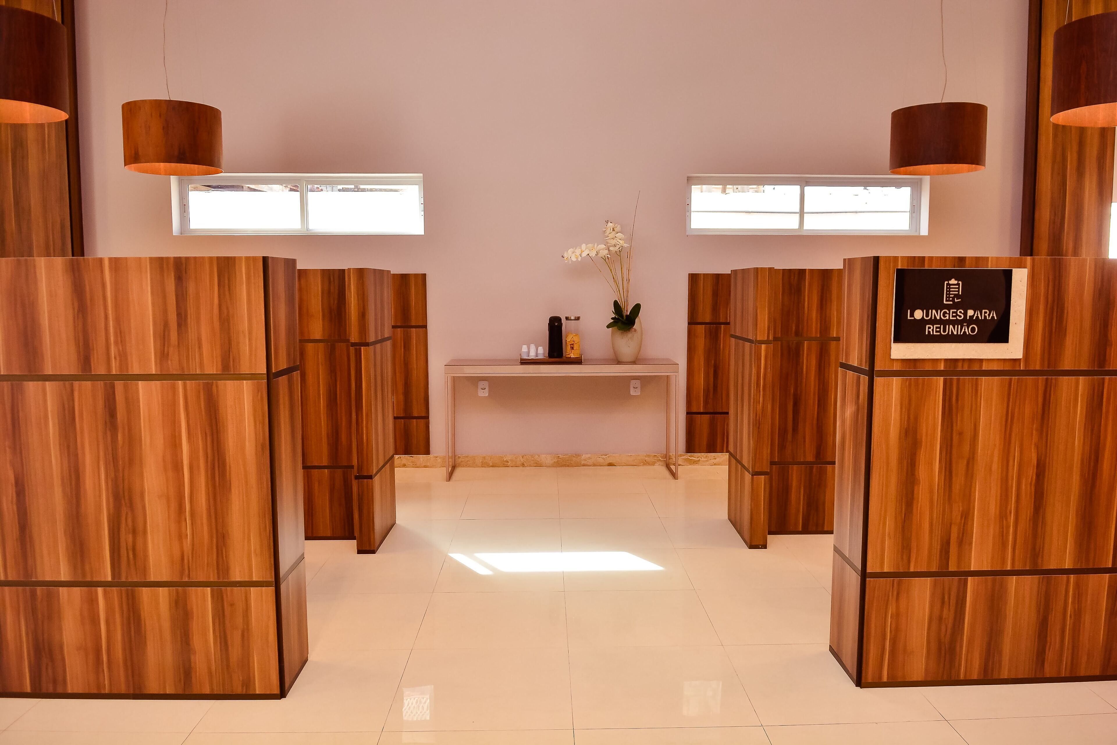 A reception area with wooden reception desks, a small counter with a vase of flowers, and a sign reading 'Lounge para reunião'.