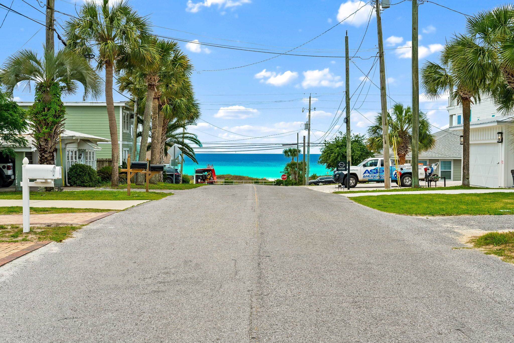 Una calle que lleva a una playa, con casas a ambos lados, palmeras y cielo despejado.