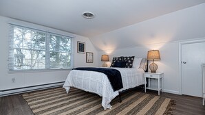 Ein Schlafzimmer mit großem Fenster, einem Bett mit weißer und dunkelblauer Bettdecke, zwei Nachttischlampen, einem kleinen weißen Nachttisch und einem dunklen Holzboden.