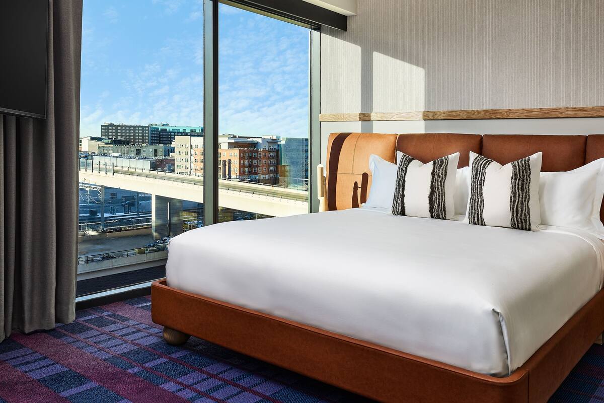 A hotel room with a large bed, a wooden headboard, a bedside table with a radio, and a view of the city through large windows.