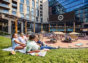 A group of people relaxing on a blanket in a grassy area with a modern building in the background.