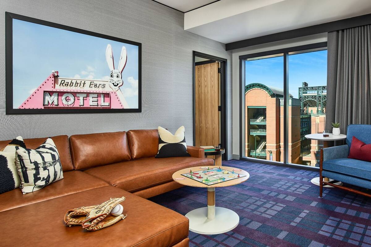 A hotel room with a brown leather sofa, a blue armchair, a small round table, and a large window offering a view of a building with a green structure.