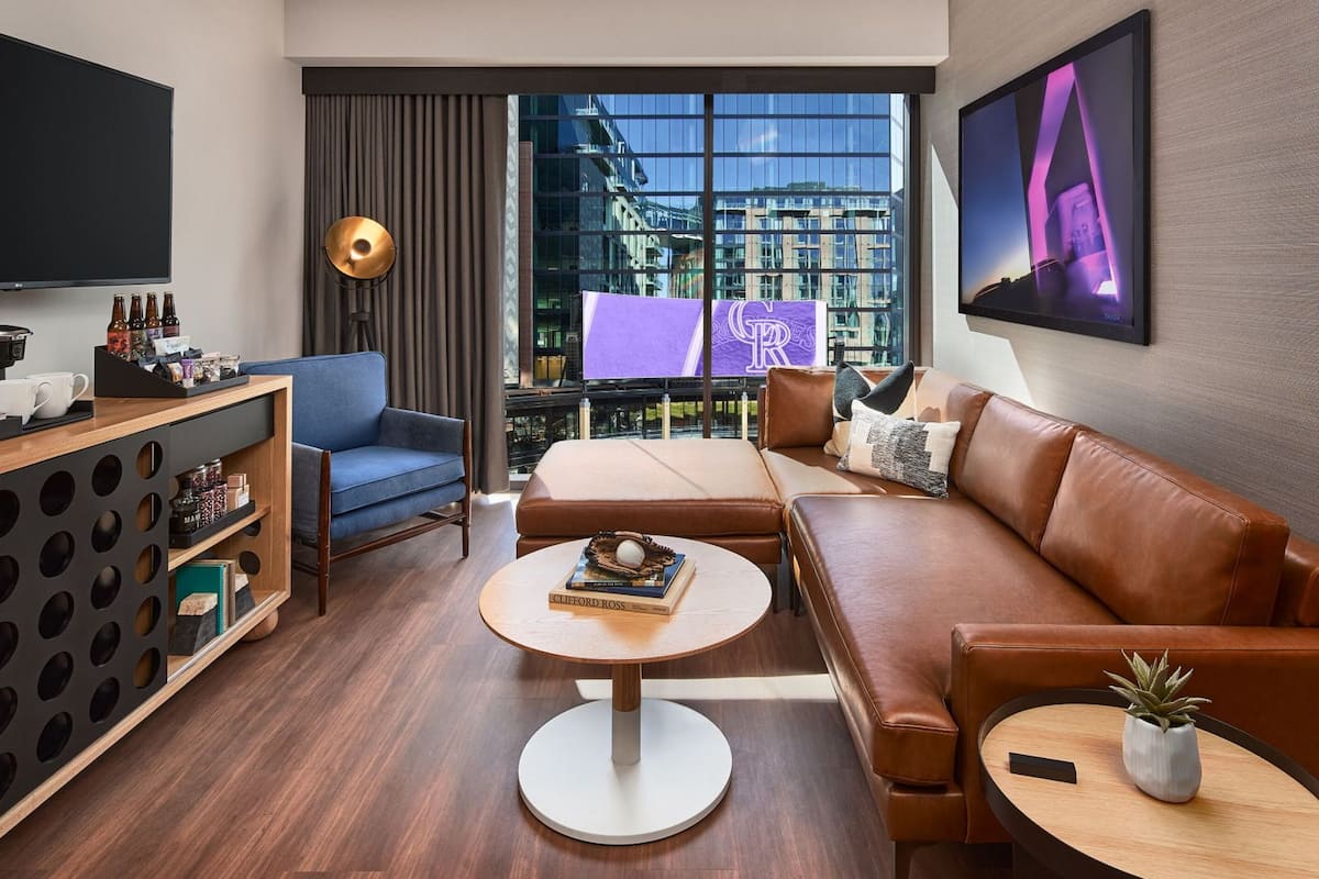 A modern hotel room with a sofa, a coffee station, a TV, and a view of the city through large windows.
