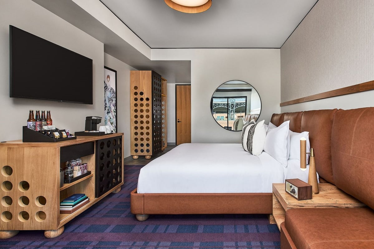 A modern hotel room with a large bed, a wooden side table, a TV, a minibar, and a view of the city through a circular window.