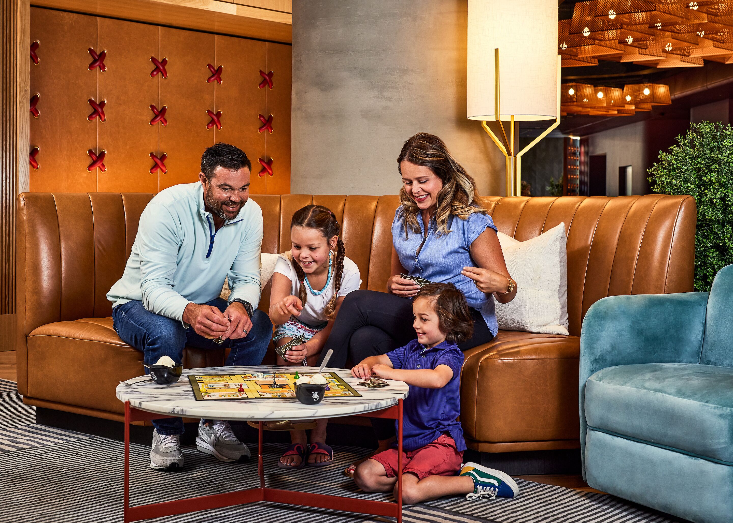 A family playing board games on a brown leather couch with a marble coffee table in a modern lobby with wooden paneling and a chandelier.