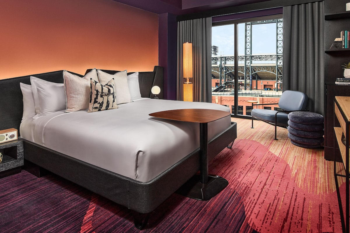 A modern hotel room with a large bed, a bedside table with a lamp, a small side table, a desk with a chair, a bookshelf, and a view of a bridge and buildings.
