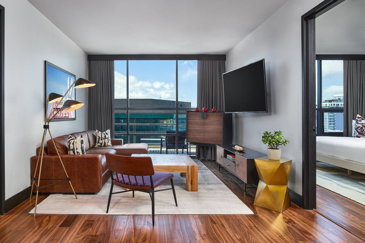 A modern hotel room with a large window, a sofa, a coffee table, a TV, and a plant.