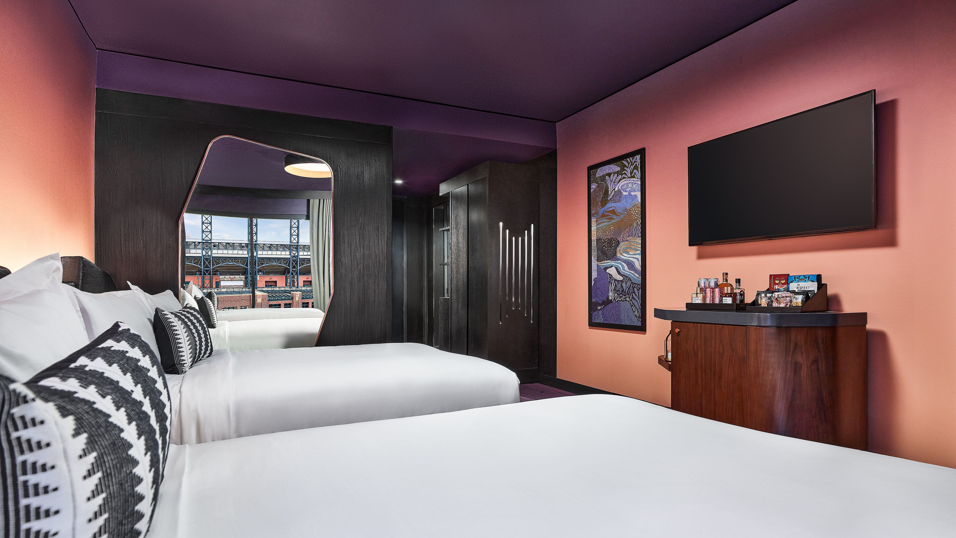 A hotel room with a bed, a desk with a flat-screen TV, a minibar, and a large window with a view of a stadium.