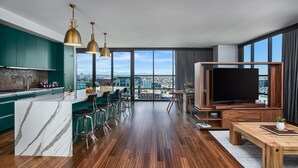 A modern kitchen with a marble countertop, a dining area with bar stools, a large flat-screen TV, and a city view through floor-to-ceiling windows. - The Rally Hotel at McGregor Square (Denver)
