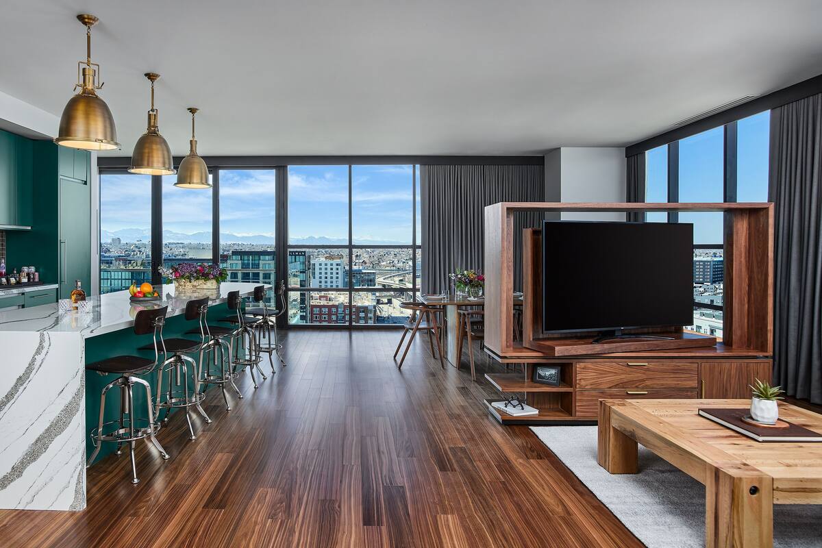 A modern kitchen with a marble countertop, a dining area with bar stools, a large flat-screen TV, and a city view through floor-to-ceiling windows.