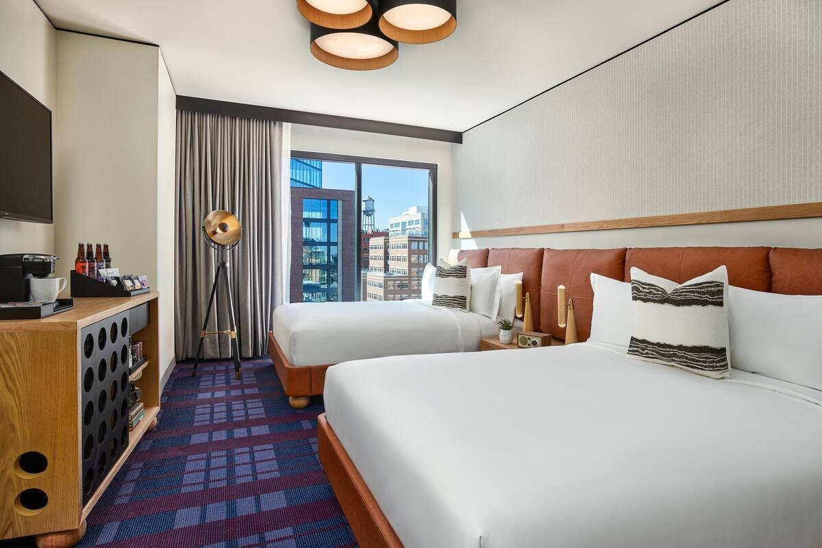 A hotel room with two beds, a desk with a coffee machine, a minibar, and a large window with a city view.