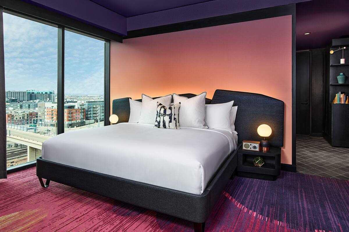 A modern hotel room with a large bed, a city view, and a bookshelf.