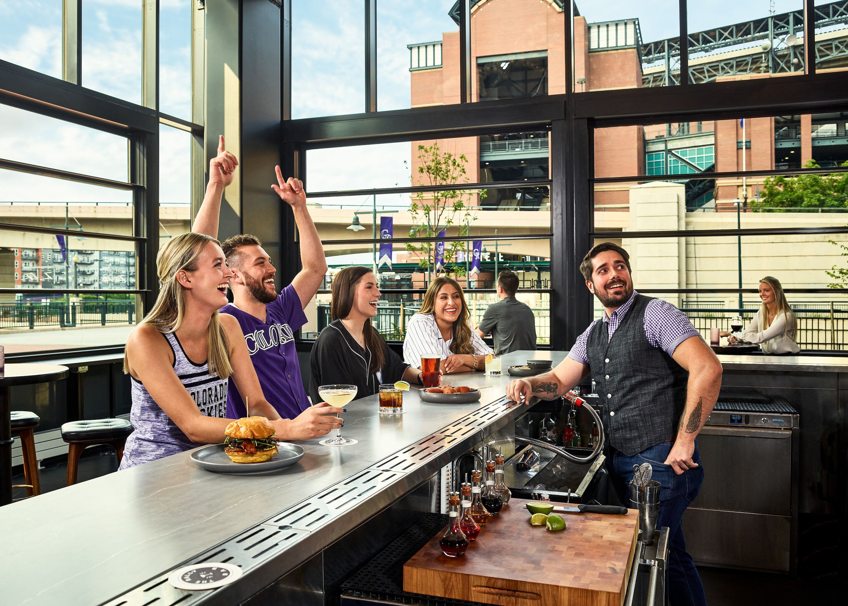 A group of people at an outdoor bar with a view of a bridge and buildings.