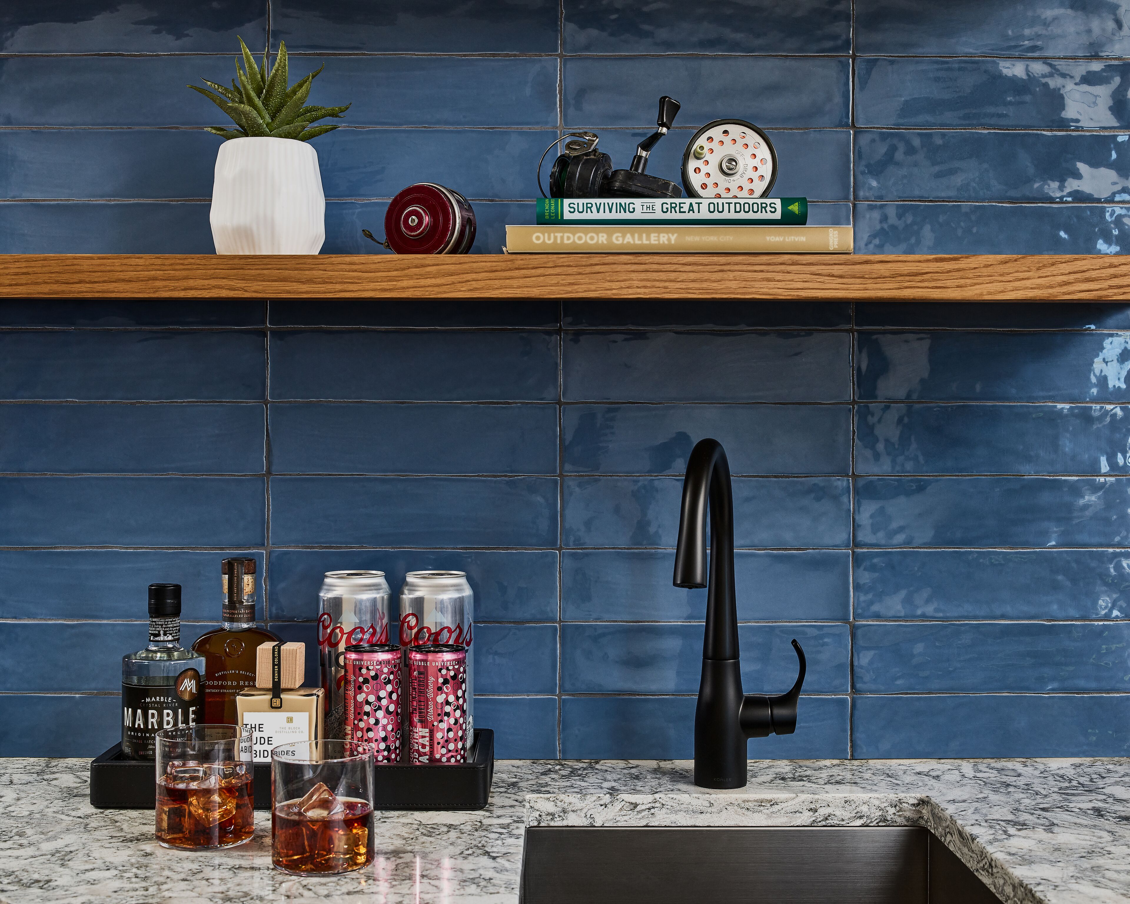 A kitchen with a blue tiled backsplash, a wooden shelf with books and a plant, a modern faucet, and a marble countertop with bottles and glasses.