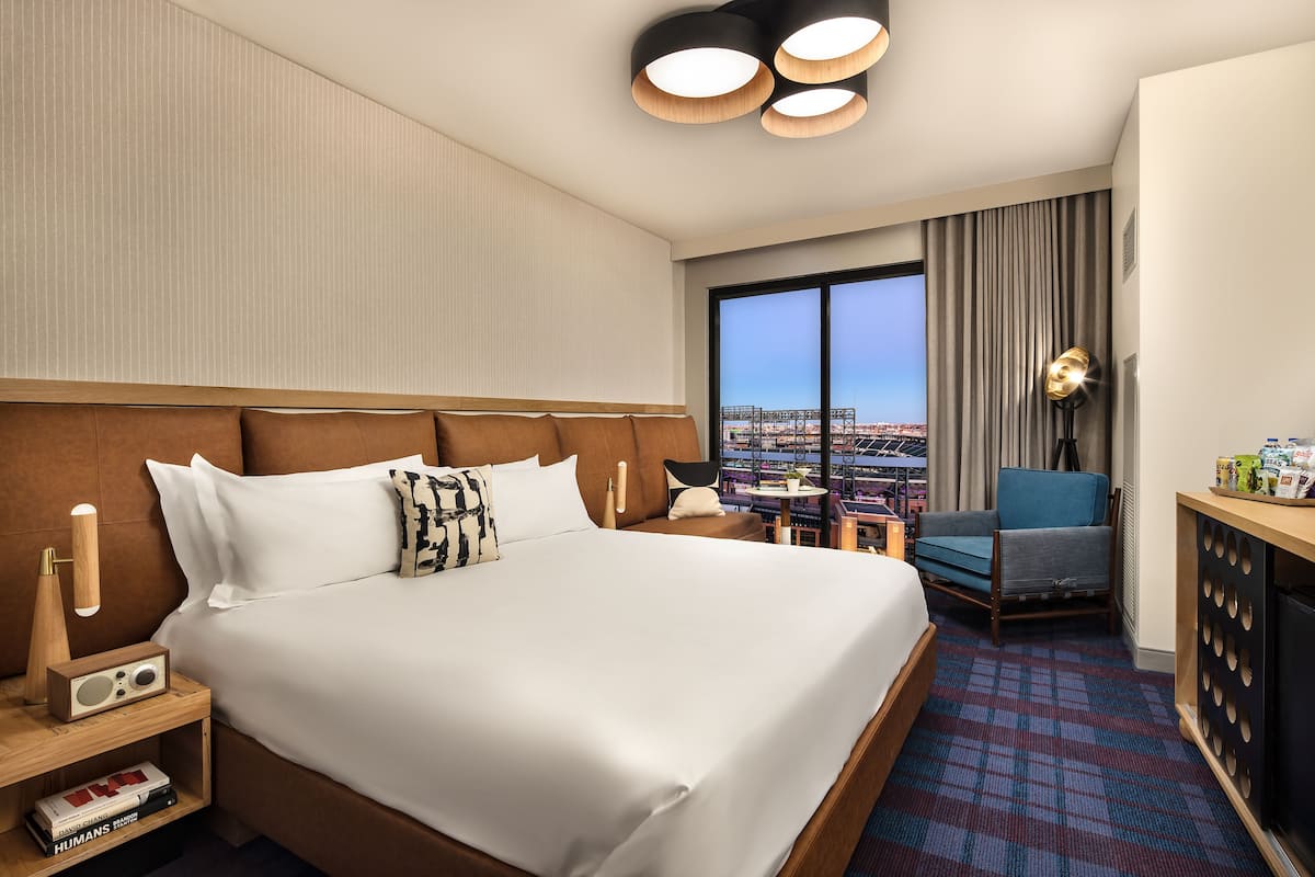 A hotel room with a large bed, a desk with a lamp, a chair, and a view of a cityscape through a large window.