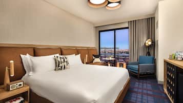 A hotel room with a large bed, a desk with a lamp, a chair, and a view of a cityscape through a large window.