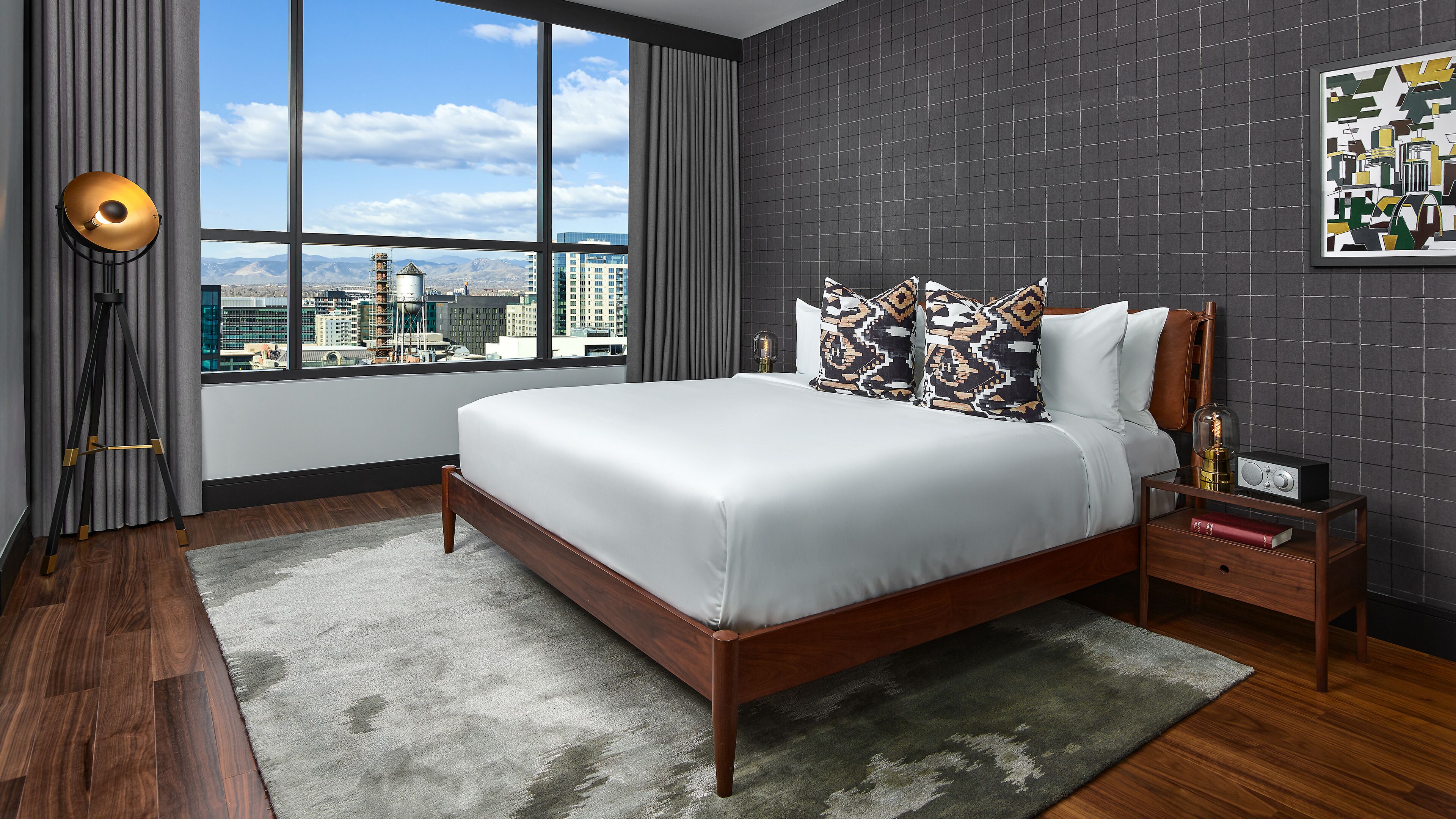 Ein modernes Schlafzimmer mit einem großen Bett, Blick auf die Stadt, einer dunkel gefliesten Wand und einem gerahmten Kunstwerk.