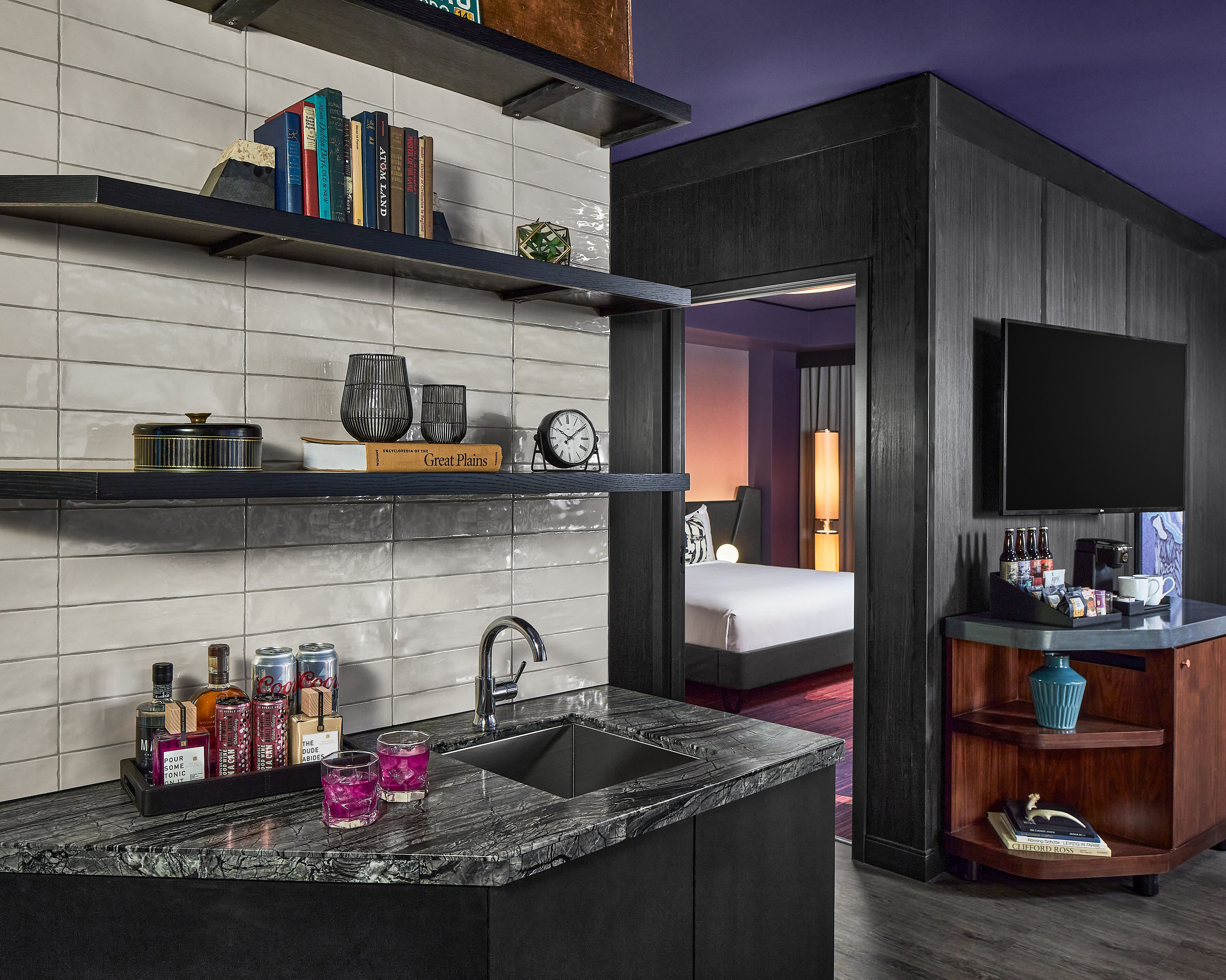 Habitació d'hotel moderna amb taulell de marbre, un rentapeus i un minibar. Hi ha llibres a una estant, un rellotge i una televisió penjada a la paret.