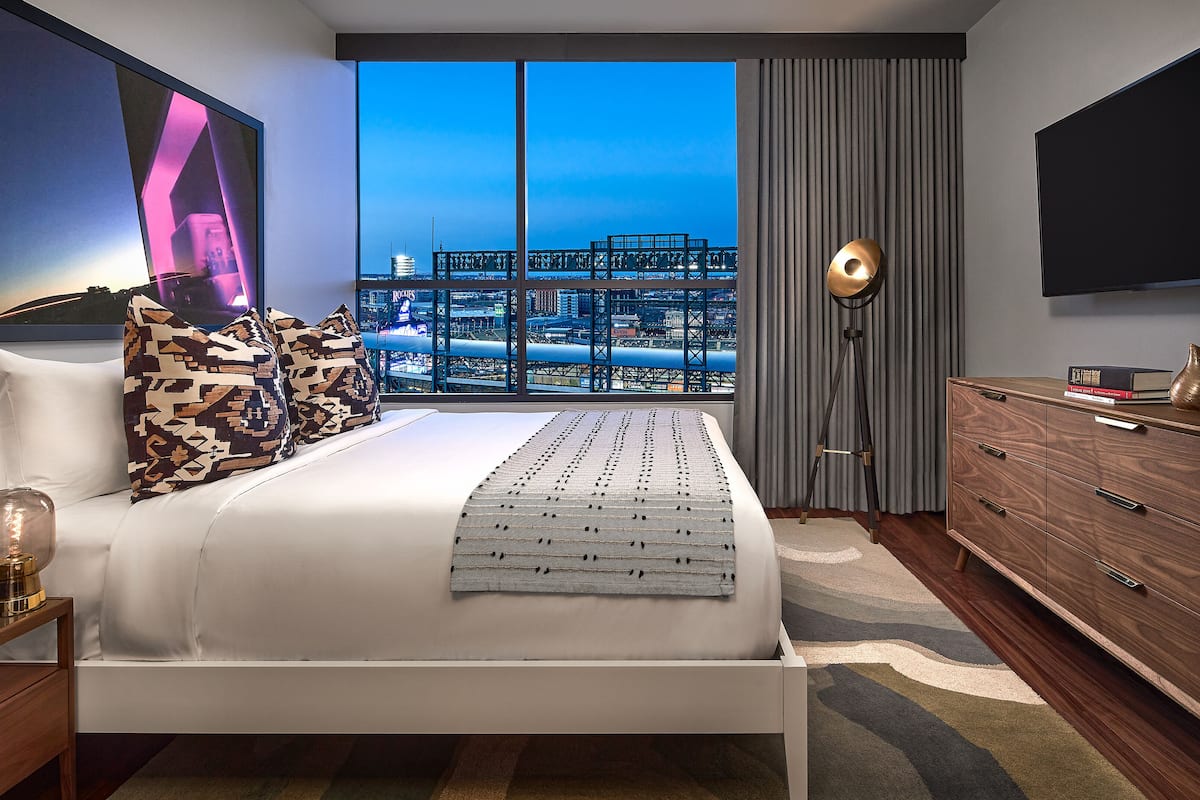 A modern bedroom with a large bed, a city view, a standing lamp, and a TV on the wall.