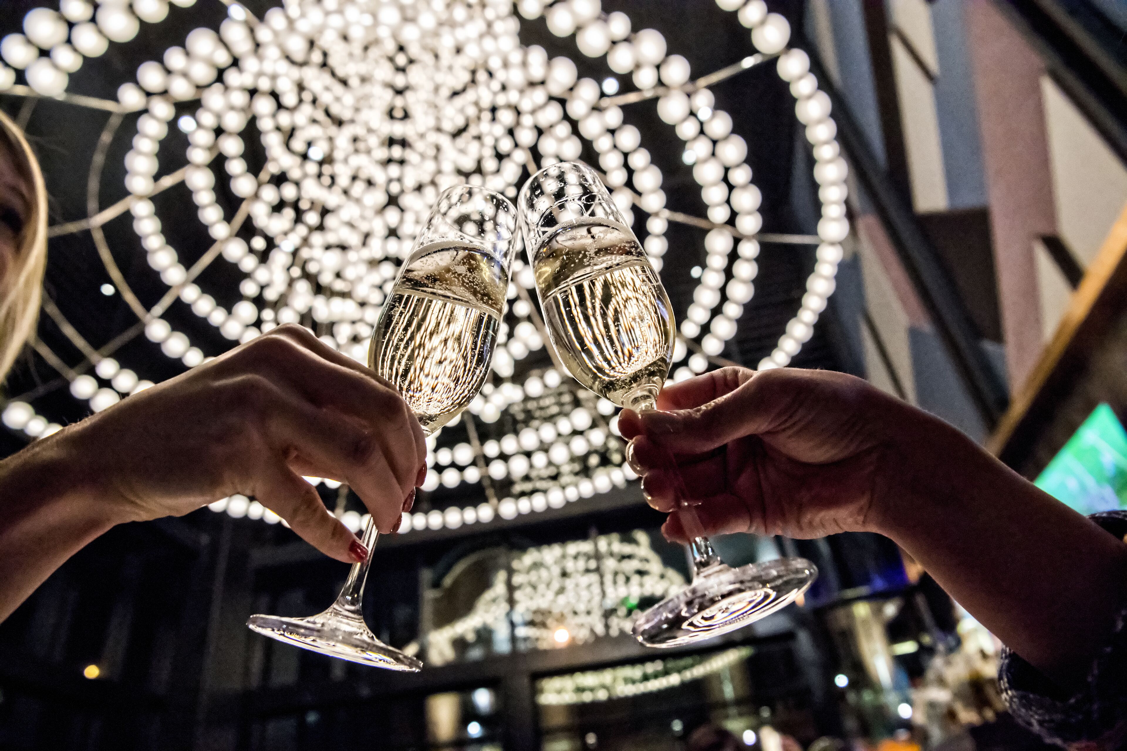 Two hands holding champagne flutes in a dimly lit setting with a circular light fixture in the background.