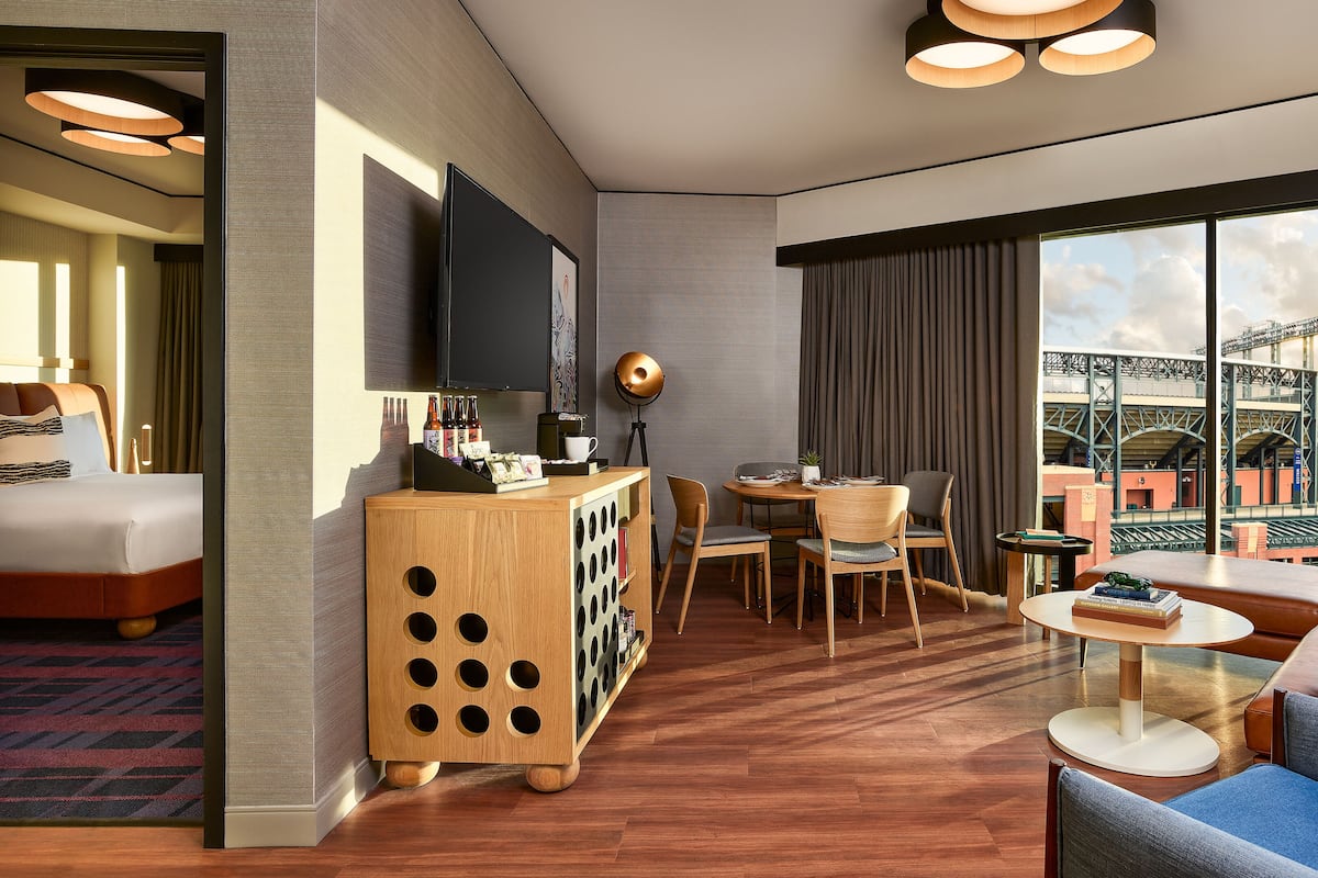 A hotel room with a bed, a wooden sideboard with bottles, a dining area with a table and chairs, and a large window with a view of a stadium.