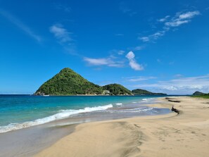 A beach with clear blue water, sandy shore, and green hills in the background.
