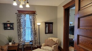 Una sala de estar acogedora con piso de madera, un tapete marrón, un sofá, un reposapiés, una silla, una mesita de noche con una lámpara, una estantería y una ventana con cortinas estampadas.