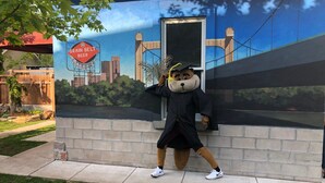 Une mascotte portant un mortier et une toge de remise de diplômes, posant devant une fresque représentant un pont et un paysage urbain.
