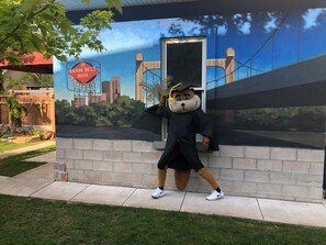 A mascot in a graduation cap and gown posing in front of a mural with a bridge and cityscape. - Enjoy this amazing home and yard as you eat, pray and fall in love with NE Mpls. (Minneapolis)