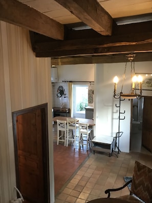 Interior rústico com vigas de madeira aparentes, área de jantar com cadeiras brancas, um lustre e cozinha visível ao fundo.