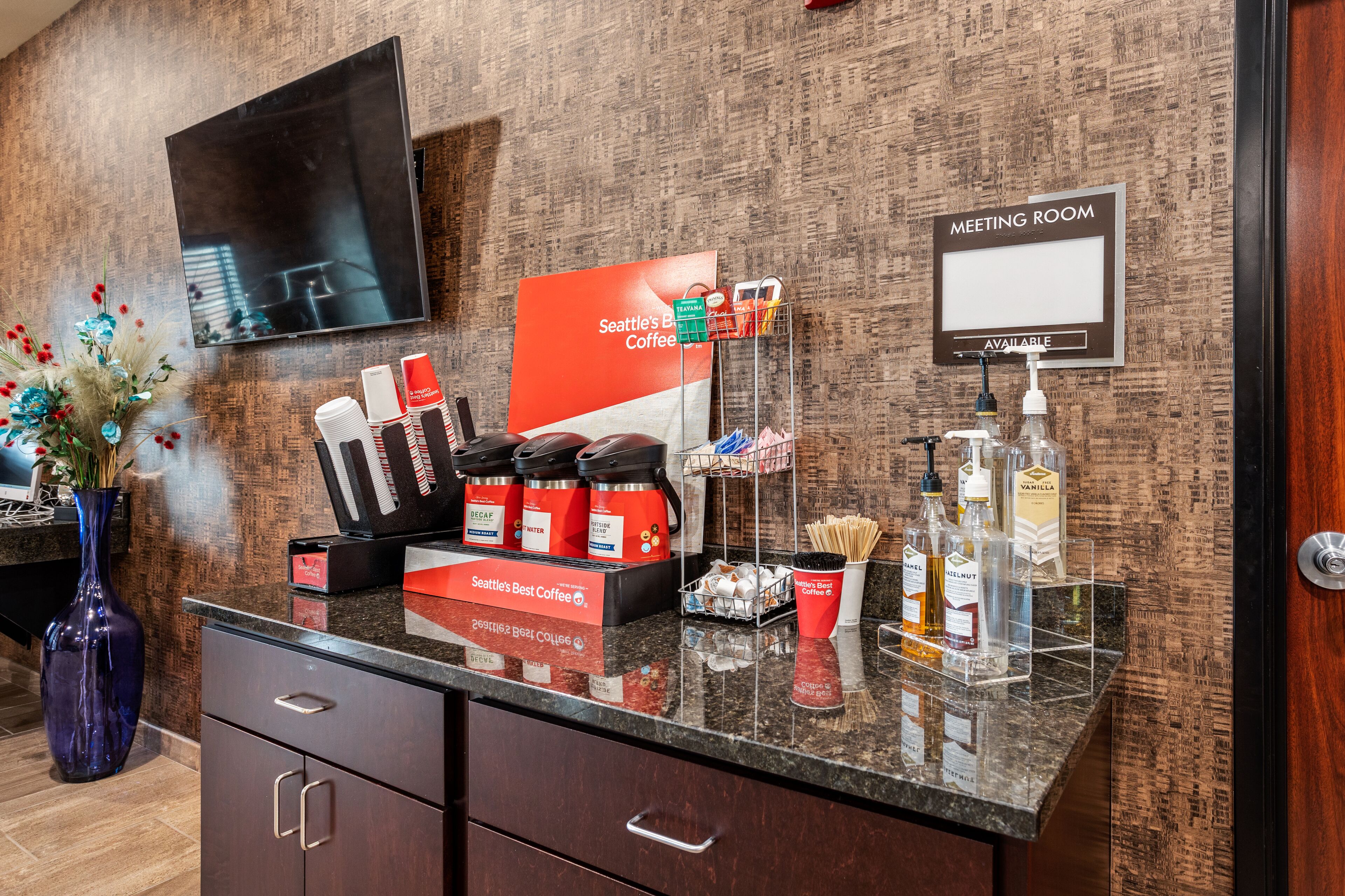 A hotel reception area with a coffee station, a meeting room sign, and a vase with flowers.