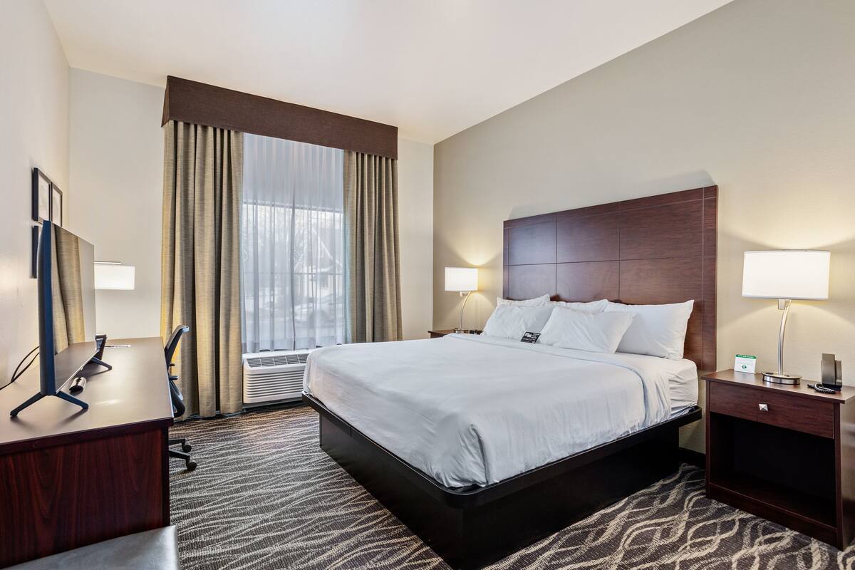 Habitación de hotel con una cama grande, un escritorio, una silla, una lámpara y una ventana con cortinas.