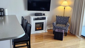 Un salon doté d’une cheminée, d’un téléviseur fixé au mur, d’un fauteuil à motifs et d’un coin repas avec une table et des chaises.